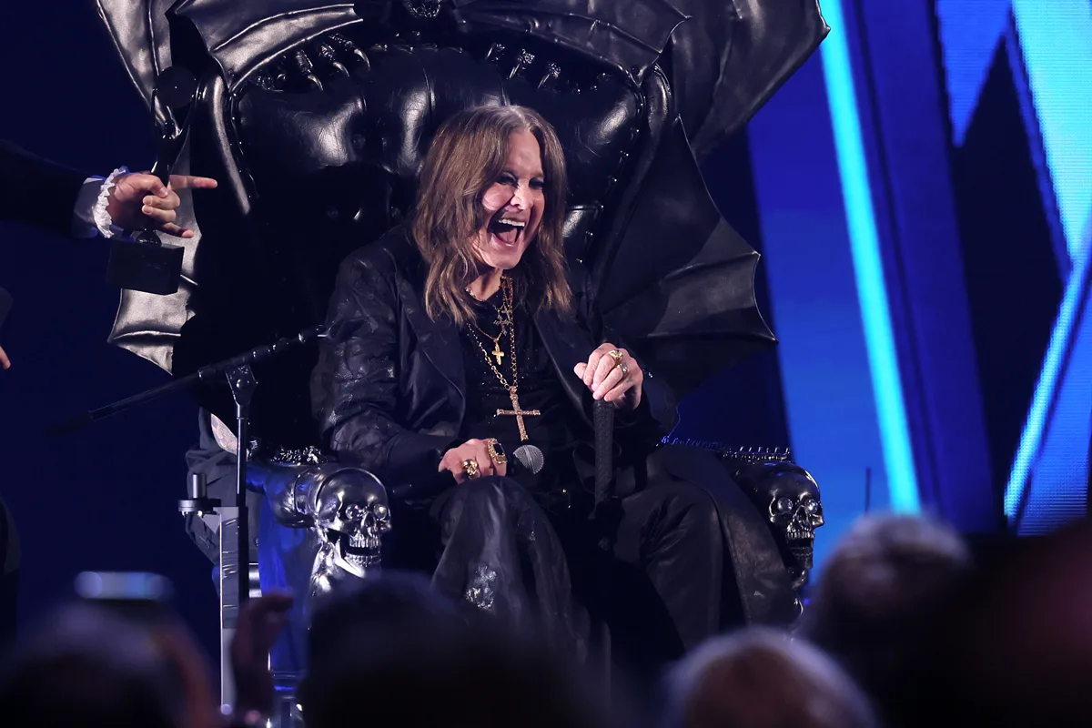 Ozzy Osbourne, a white man with long dark hair and gothic clothing, sits on a black throne-style chair, with blue lighting behind and a crowd in front of him